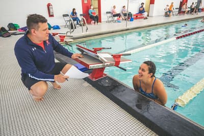 Coach on deck talking with a swimmer at the wall, both smiling — the mentorship relationship
