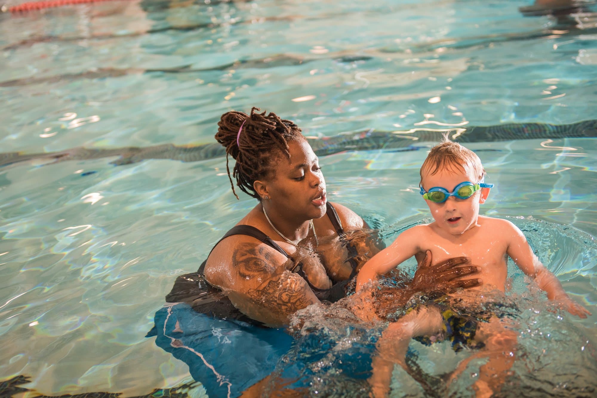A coach supporting a toddler in goggles as he kicks through the water